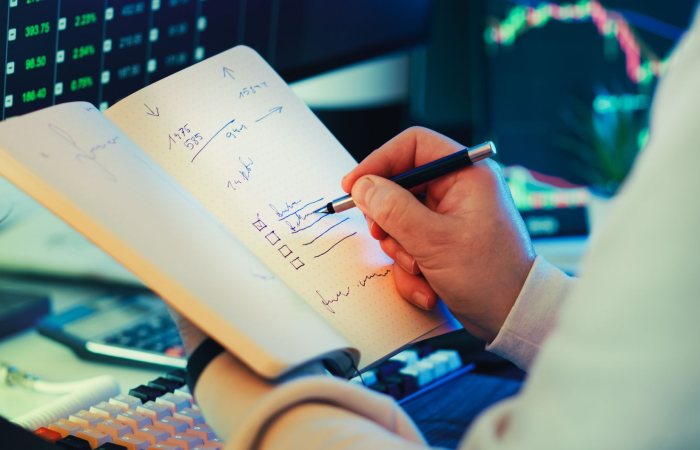 A businessman writes notes in a notebook while analyzing stock market data on a computer screen.