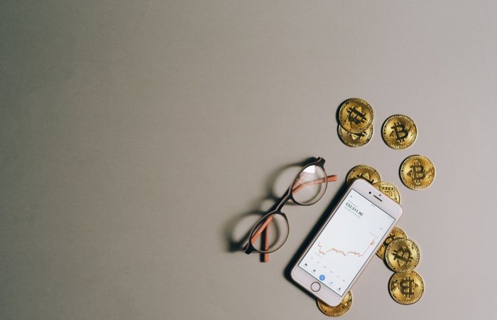 Flat lay of Bitcoin coins, smartphone with graph, and glasses on gray surface.