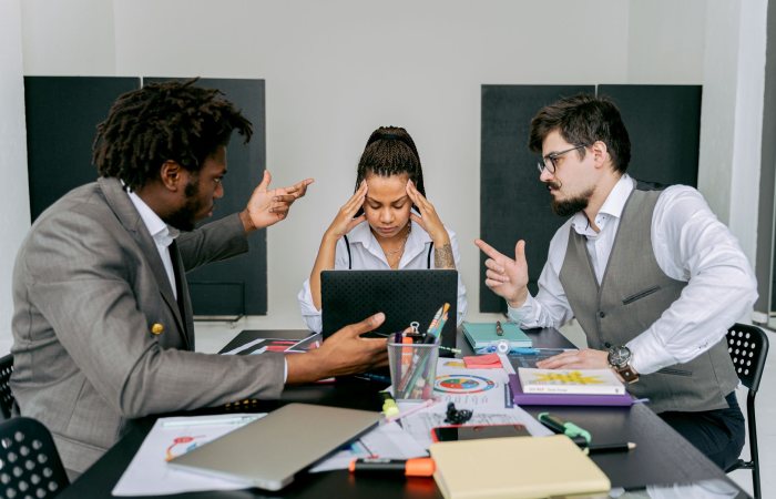 Two men argue while a woman looks frustrated at a laptop in an office environment.