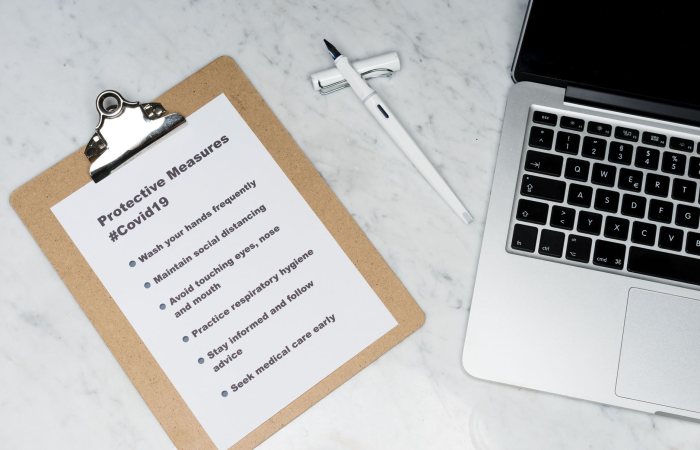 Flat lay of a clipboard listing Covid-19 safety measures beside a laptop on a marble surface.