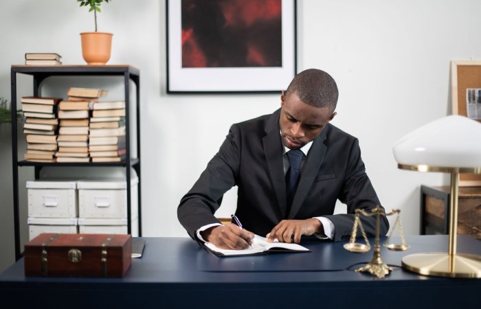 Businessman in formal suit writing notes at office desk with books and scales of justice.