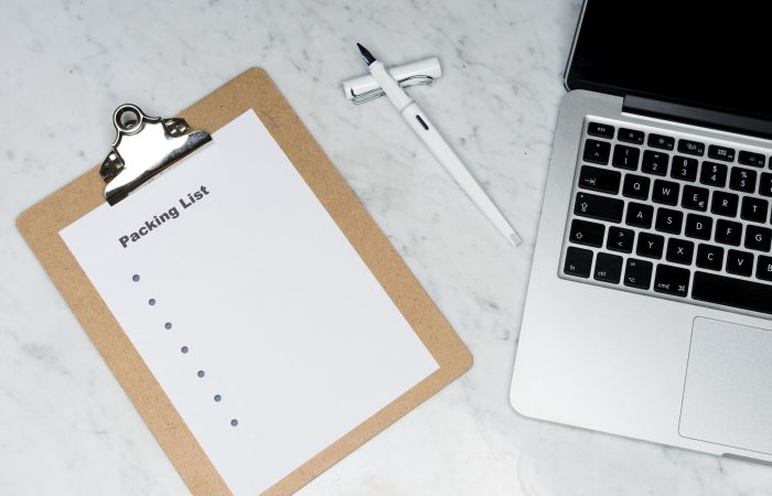 Flat lay of a packing list clipboard with a laptop and pen on a marble desk.